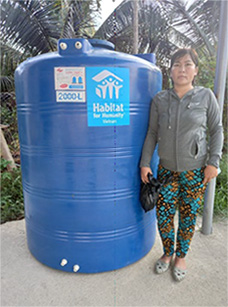 「Habitat for Humanity Vietnam」のロゴが入った2000リットルの青い貯水タンクの横に立つ女性。