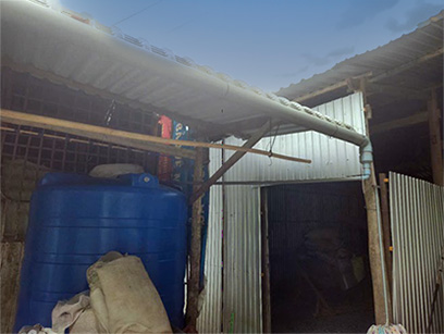 屋根の雨どいからパイプでつながれた青い貯水タンクが、簡易建物のそばに設置されている。