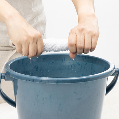 青いバケツの上で、両手で濡れた白い布を絞って水を落としている様子。布から水滴が滴り落ちている。