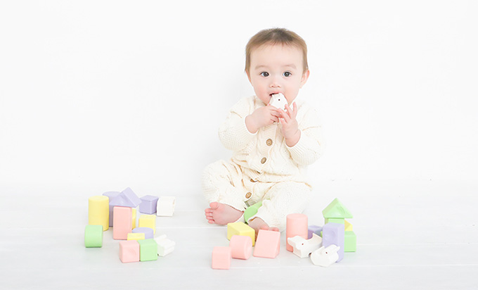 ライスレジン製の玩具で遊ぶ乳児のイメージ。淡いパステルカラーの積み木や動物型パーツが床に並び、原料となるお米の種類や状態によって生まれる自然な色合いの違いが表現されている。人工的に均一化されたプラスチックにはない、自然由来のやさしい風合いを伝えている。