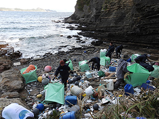 海に面した岩場の海岸で、大量の漂着ごみを回収するツアー参加者たち。プラスチック容器やブイ、発泡スチロールなどが散乱しており、作業員たちは緑色の大型袋に手分けして回収作業を行っている。背後には岩壁と波打ち際が広がる自然環境。