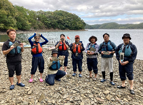 海辺立つツアー参加者のグループ。全員がライフジャケットを着用しており、手には岩の破片のようなものを持って掲げている。