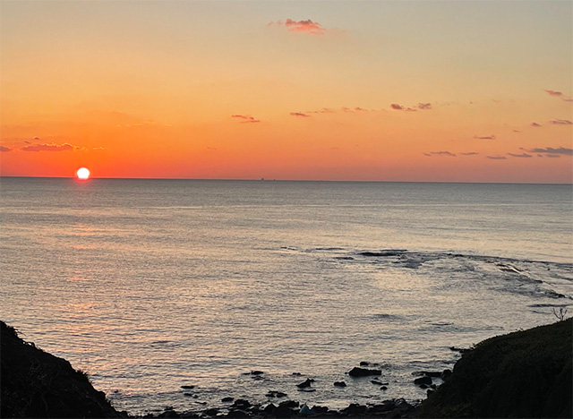 海に沈む夕日が水平線近くに輝く、美しい夕景の写真。空はオレンジから紫がかったグラデーションに染まり、穏やかな海面に反射する光が幻想的な雰囲気を醸し出している。手前には岩場と草地のシルエットが見える。