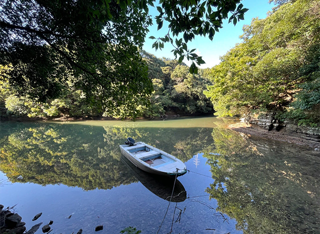 緑に囲まれた静かな入江に一艘の小型ボートがロープで係留されて浮かんでいる。水面には青空と木々が美しく反射しており、自然の静けさと透明感あふれる景色が広がる。