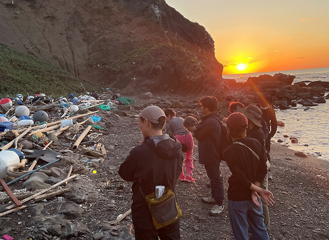 夕日が沈む海岸で、漂着ごみの山を前に立ち尽くすツアー参加者のグループ。プラスチックごみや流木、発泡スチロールなどが岩場に散乱し、参加者たちは静かにその現状を見つめている。背後には夕焼けに染まる空と海が広がる。