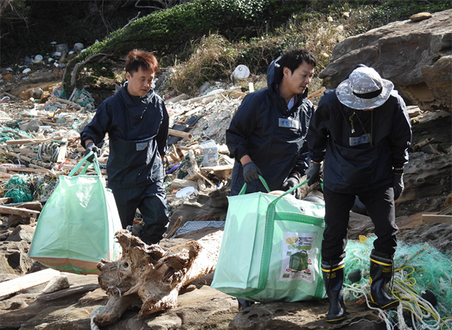 海岸に散乱した大量の漂着ごみの中で、3人のツアー参加者が緑色の回収用バッグを持って清掃作業を行っている様子。背景にはロープや木材、プラスチックごみが広がっており、清掃活動の大変さがうかがえる。作業員は手袋や長靴を着用し、真剣に作業している。