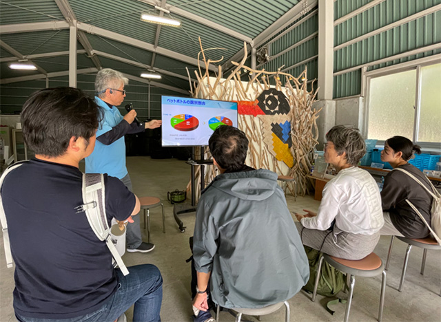 屋内施設で、講師がモニターを使って「ペットボトルの国別割合」に関するプレゼンテーションを行っている様子。モニターには円グラフが表示され、数名の参加者が椅子に座って真剣に話を聞いている。背景には流木を組んだ装飾やカラフルな壁アートが見える。