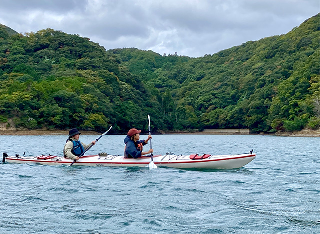 青く穏やかな水面を、2人乗りのカヤックに乗って漕ぐツアー参加者。ライフジャケットを着用し、パドルを使って前進している。背景には緑豊かな山々が広がっている。