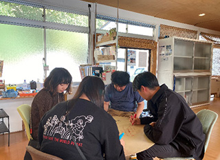 明るい室内で、4人の参加者が円卓を囲み、クラフト紙のような大きな丸い紙にカラーペンで書き込みをしている様子。ワークショップを真剣に取り組んでいる。