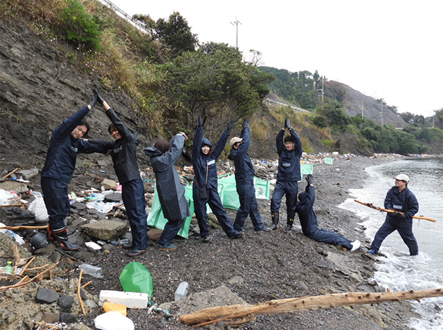 海岸の漂着ごみ清掃活動中、作業服姿の8人がごみの中でポーズを取りながら笑顔を見せている。うち7人は「ASKUL」と読めるような人文字を作っており、1人は木の棒を持ってアスクルのキャラクターに扮している。背景には回収用の緑色の袋と散乱するプラスチックごみが見える。