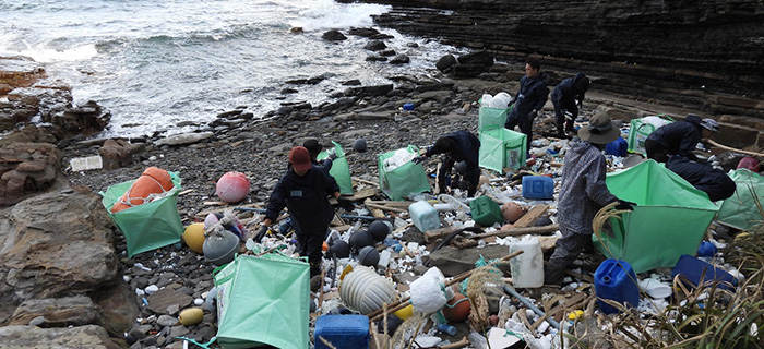 海に面した岩場の海岸で、大量の漂着ごみを回収するツアー参加者たち。プラスチック容器やブイ、発泡スチロールなどが散乱しており、作業員たちは緑色の大型袋に手分けして回収作業を行っている。背後には岩壁と波打ち際が広がる自然環境。